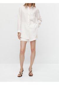 Leinen-Hemd in hellem Creme mit langen Ärmeln, kleiner Brusttasche und abgerundetem Saum, kombiniert mit cremefarbenen Shorts und schwarzen Sandalen.