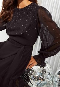 Svart klänning med genomskinliga puffärmar, prydd med silverfärgade rhinestones. Midjan har ett knytband, tillsammans med reflekterande diskokulor.