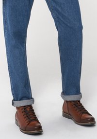 Brązowe skórzane botki na kostkę z ciemnymi sznurówkami, w połączeniu z ciemnoniebieskimi dżinsami z podwiniętymi nogawkami. Powierzchnia jest gładka z subtelnym połyskiem.