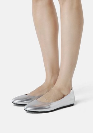 Person wearing shiny silver ballet flats while standing on a white surface, showing lower legs and feet.