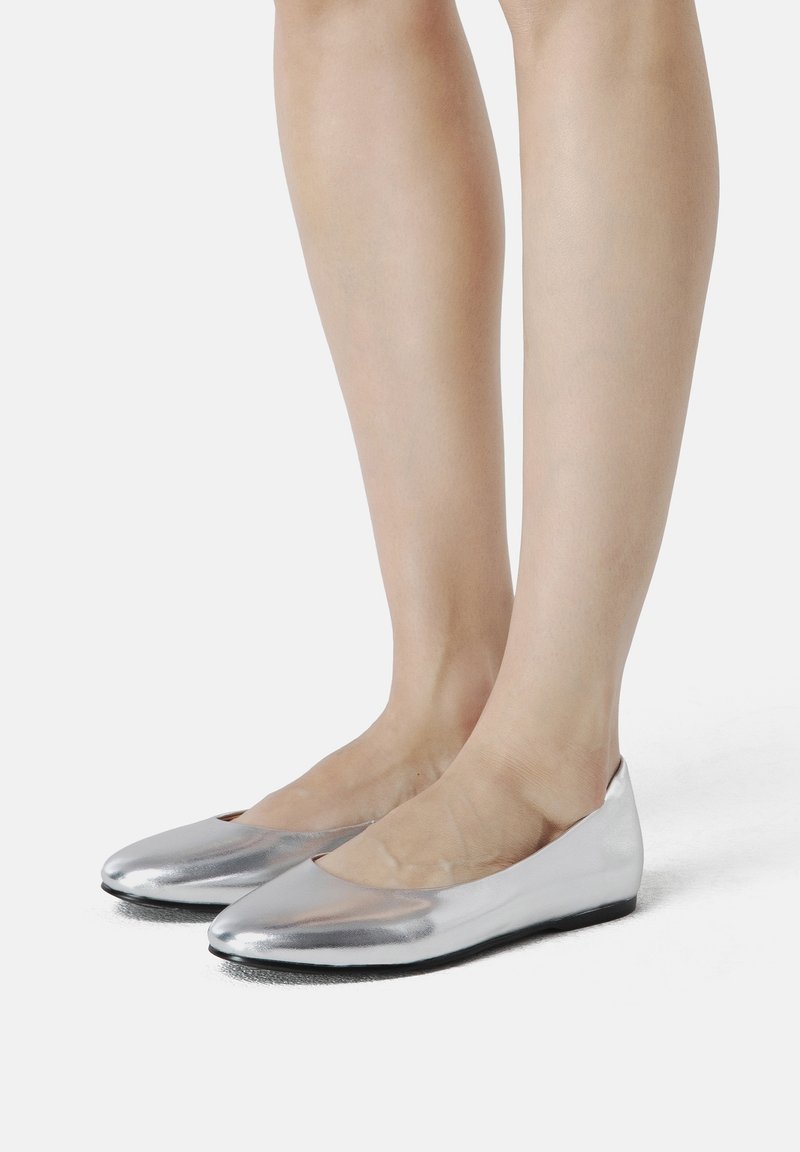 Personne portant des ballerines argentées brillantes debout sur une surface blanche, montrant les jambes et les pieds.
