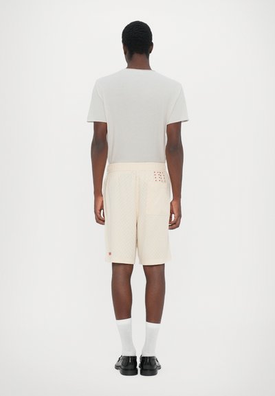 Heren lichte beige shorts met subtiel ruitpatroon, elastische tailleband, zijzak en rode accentdetails. Gecombineerd met een grijs T-shirt met korte mouwen.