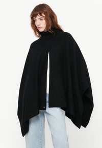 Musta neulottu poncho, jossa on korkea kaulus, suvaitseva istuvuus ja epäsymmetrinen helma, yhdistettynä vaaleansinihin farkkuihin ja valkoiseen paitaan.