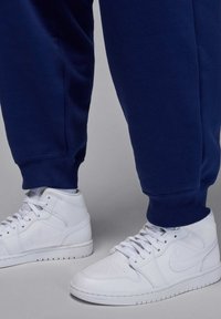 Person iført hvide Nike høje sneakers kombineret med marineblå opbrettede joggingbukser, stående på en lysegrå overflade.