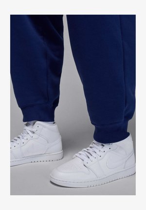 Henkilö, jolla on valkoiset Nike-tennarit ja tummansininen resorilla varustetut collegehousut, seisoo vaalean harmaalla pinnalla.