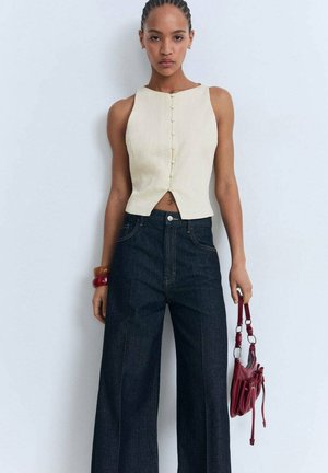 Top sin mangas de color crema con botones en la parte delantera, combinado con pantalones de mezclilla oscuros de pierna ancha y cintura alta, y un pequeño bolso rojo.