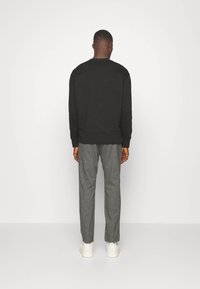 Homme debout, de dos, portant un haut noir à manches longues, un pantalon gris et des baskets blanches, sur un fond blanc uni.