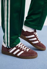 Sneakers in camoscio marrone scuro con tre strisce bianche, lacci bianchi e suola in gomma marrone, abbinate a pantaloni da jogging verdi con strisce bianche laterali.