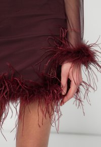 Bordo haljina od teksturiranog materijala, sa dugim resama od perja na rubu i rukavima. Ruka je vidljiva i drži haljinu.