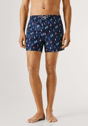 Męski tors i nogi ubrany w granatowe kąpielówki z wielokolorowymi wzorami żaglówek, stojący boso na jednolitym tle.