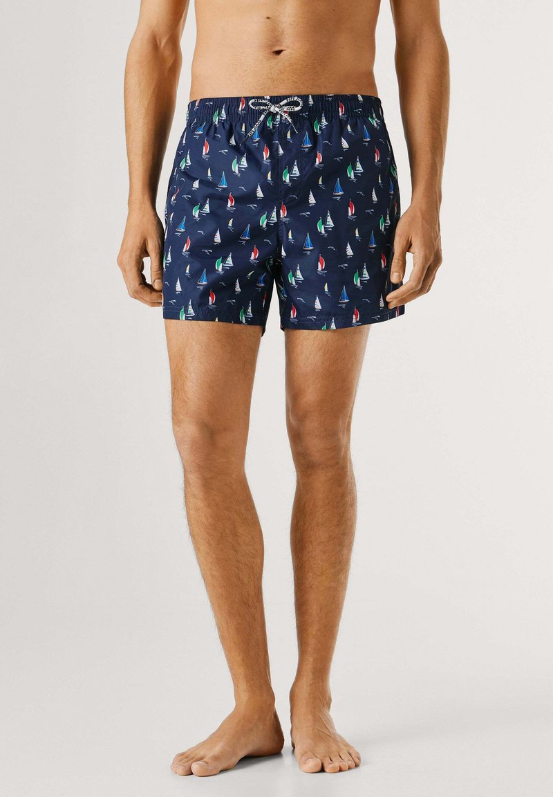 Meespealne torso ja jalad, kellel on seljas tumesinised ujumispüksid, millel on mitmevärvilised purjekate mustrid, seisab paljajalu ühevärvilise tausta ees.