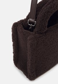 Sac à main en tissu teddy texturé marron foncé avec une forme rectangulaire et une sangle en cuir foncé fixée par des ferrures argentées.