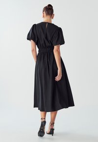 Črna midi obleka s puhovimi rokavi, elastičnim pasom in ključavnico na hrbtni strani. Gladko blago in tekoč dizajn. Obuta v petke.