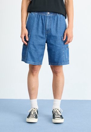 Blauwe denim cargoshorts met een elastische tailleband, zijzakken en een relaxte pasvorm. Draagt zwarte sneakers en witte sokken.