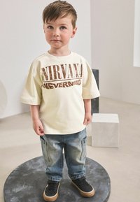 T-shirt crème avec impression marron "NIRVANA NEVERMIND", associée à un pantalon en denim bleu clair et des baskets noires avec semelles en caoutchouc.