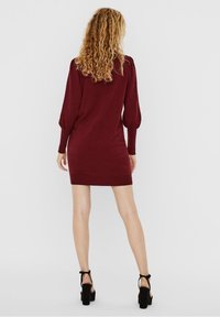 Femme aux cheveux blonds bouclés, portant une robe courte à manches longues bordeaux et des sandales à talons noirs, se tenant de dos sur un fond uni.