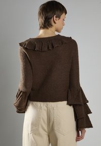 Brauner Strickpullover mit Rüschensaum am Kragen und an den Ärmeln, aus strukturiertem Stoff. Getragen mit cremefarbenen Hosen. Rückansicht gezeigt.