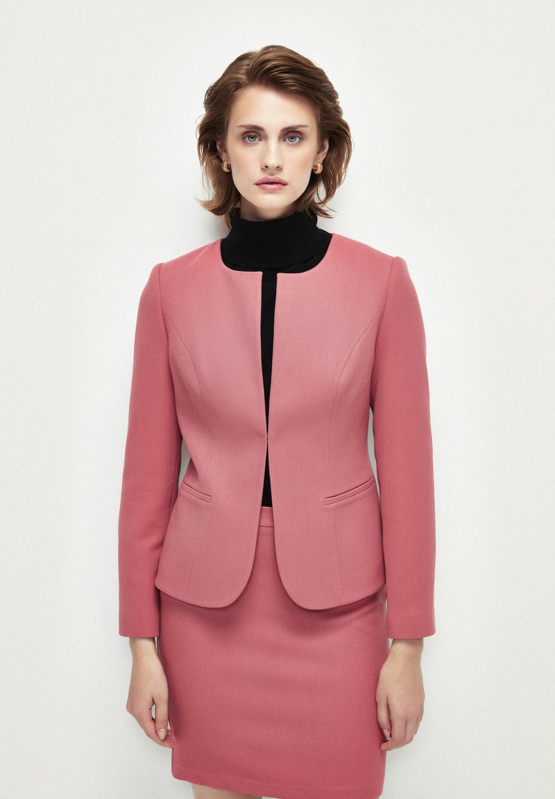 Blazer tailored color rosa pallido con scollatura rotonda, due tasche frontali e design aderente, abbinato a una gonna a matita coordinata. Maglione nero a collo alto sotto.