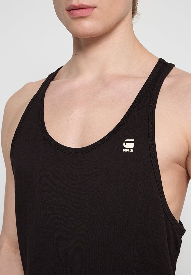 Close-up van een persoon die een zwart mouwloos sporttopje draagt met een klein wit "RAW" logo op de linkerkant van de borst.