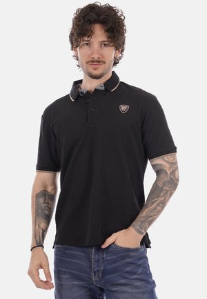 Uomo con capelli ricci che indossa una polo nera e jeans blu, mostrando tatuaggi dettagliati su entrambi gli avambracci, in piedi con una mano in tasca.