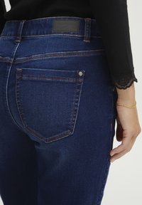 Temno modri jeans iz denima, ki se ponašajo s klasičnim dizajnom s petimi žepi, vidno šivom v oranžni barvi in teksturirano usnjeno oznako blagovne znamke na pasu.