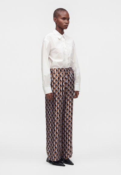 Personne portant une chemise blanche boutonnée et un pantalon à jambes larges avec un motif triangulaire géométrique marron et noir, debout devant un fond uni.