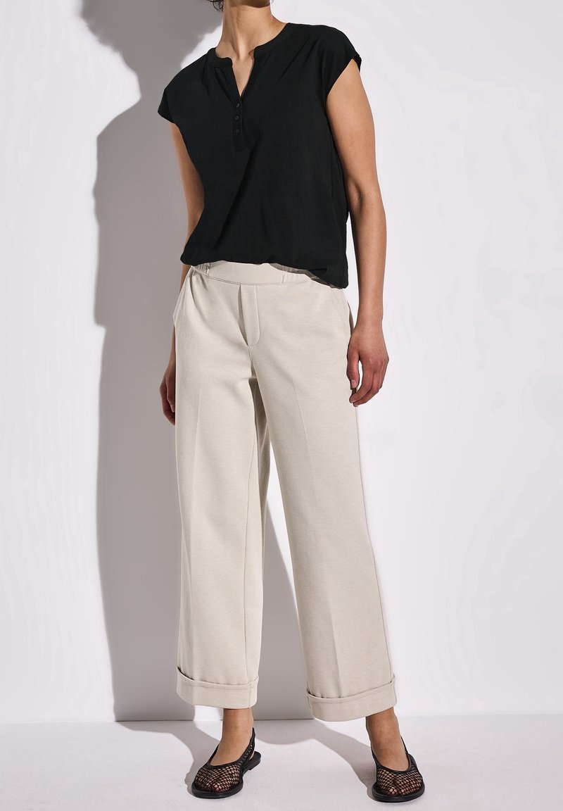 Černý krátký rukávový top s knoflíkovým výstřihem, v kombinaci se smetanovými kalhotami širokého střihu s ohrnutými manžetami a jemnými záhyby, obutý s černými balerínami.