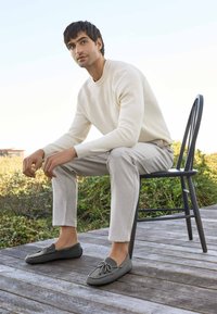 Graue Mokassinsocken mit einer Schnürdetails, getragen mit hellbeigen strukturierten Hosen und einem cremefarbenen gerippten Pullover, sitzen auf einem schwarzen Stuhl.