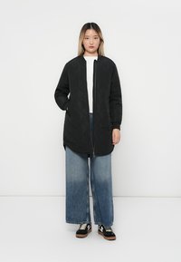 Black quilted long jacket with a zip front, round neckline, and side pockets, paired with light blue wide-leg jeans and black sneakers.