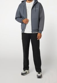 Sudadera gris con cremallera y logo, camiseta blanca debajo y pantalones negros. Zapatillas gris y blancas con detalles en negro completan el atuendo.
