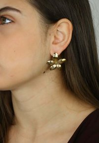 Złote kolczyki w kształcie kwiatu z teksturowanymi płatkami i centralnym detalem, noszone na płatku ucha na neutralnym tle.