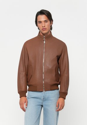 Ung mand, der står med fronten mod kameraet iført en brun lynlås læderjakke med trykknapkrave og lyseblå jeans, mod en simpel hvid baggrund.