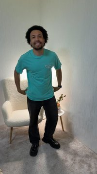 Homme souriant, portant un t-shirt bleu sarcelle et un pantalon noir, debout dans une pièce moquettée près d'un fauteuil blanc et d'une petite table ronde avec des livres et une plante.