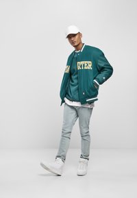 Teal varsityjack met gele letters, laagje over een teal shirt, lichtblauwe jeans en witte sneakers. Een witte pet maakt de look compleet.