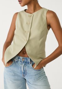 Hellgrün ärmellose Weste mit taillierter Passform, ausgestattet mit fünf beigen Knöpfen und seitlichen Taschen, kombiniert mit hellblauen Jeans.