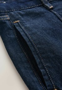 Dunkelblauer Denim-Stoff mit glatter Textur, der sichtbare Nähdetails in Hellbraun aufweist. Enthält eine Öffnung für die Vordertasche.