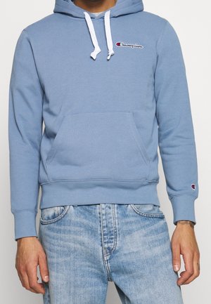 Mann trägt hellblauen Champion-Hoodie mit weißen Kordeln und Fronttasche, kombiniert mit hellblauen, ausgewaschenen Jeans.
