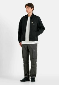Giacca nera con tasca centrale con zip, indossata su una maglietta grigia. Abbinata a pantaloni cargo scuri con multiple tasche laterali. Sneakers nere.