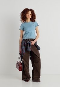 Γυναίκα με σγουρά μαλλιά, ντυμένη με ανοιχτό μπλε μπλουζάκι και καφέ φαρδιά παντελόνια, κρατώντας ένα κόκκινο τσαντάκι, με ένα καρό πουκάμισο δεμένο γύρω από τη μέση της.