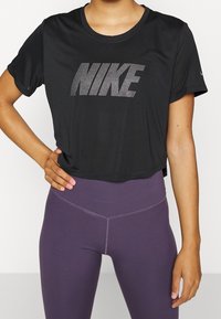 Nainen, joka on pukeutunut mustaan lyhytvartiseen Nike-t-paitaan, jossa on valkoinen logo, ja korkeavyötäröisiin violetteihin legginsseihin, käsi lantion päällä.