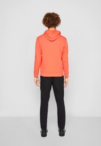 Persona con cabello rizado, vistiendo un sudadera roja con capucha, pantalones negros y zapatos negros, de pie frente a una pared blanca.