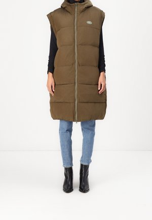 Modelka w oliwkowej bezrękawniku pikowanym bez rękawów z przednim zamkiem błyskawicznym, jasnoniebieskich dżinsach, czarnej bluzce z długim rękawem i czarnych botkach do kostki.