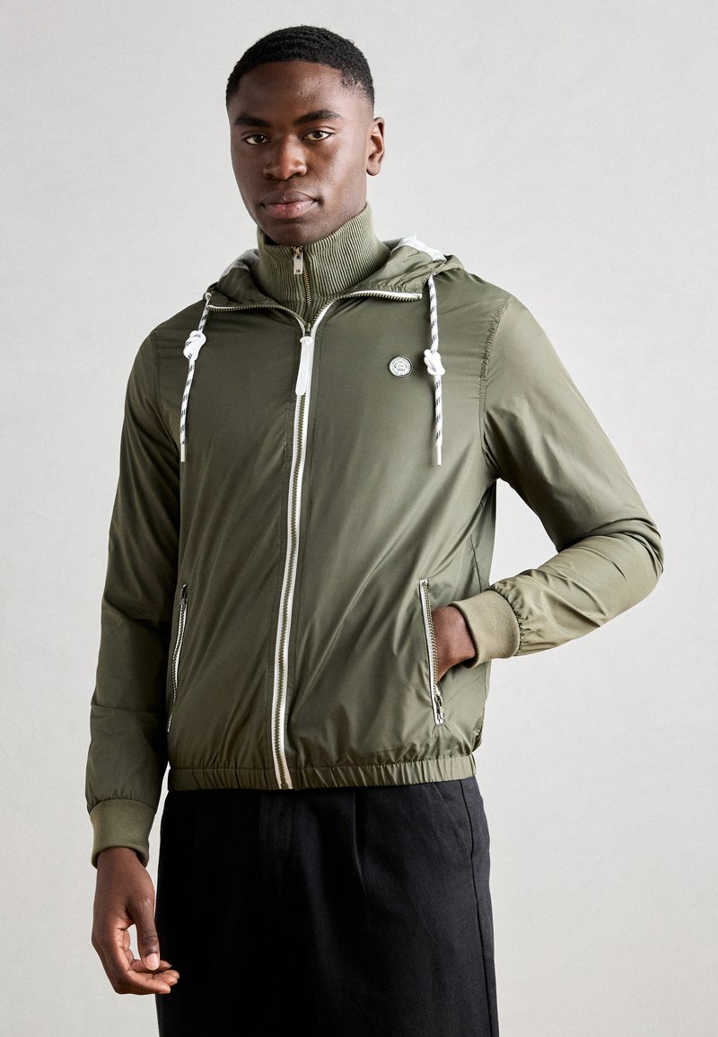 Olivegrüne leichte Jacke mit Kordelzugkapuze, frontalem Reißverschluss, Seitentaschen und gerippten Akzenten. Texturierten Stoff mit elastischen Bündchen.