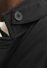 Detaliu al gulerului unei jachete negre, prezentând un material texturat, un button cu patru găuri și o cusătură precisă pe margini pentru o finisare curată.