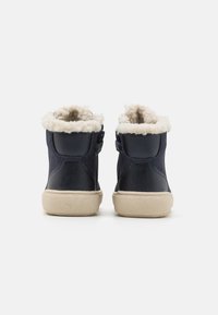 Botas de tobillo de cuero marino con forro de piel de oveja beige, suelas de goma planas y correas de velcro. El detalle de costura lateral añade textura.