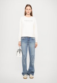 Felpa color crema chiaro con logo "Armani Exchange", jeans blu con motivo a X sulla parte anteriore e borsa bianca. Scarpe casual completano il look.