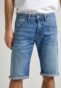 Pantalones cortos de mezclilla en azul claro, con dobladillos enrollados, diseño de cinco bolsillos y cierre de botón de metal. La textura desgastada añade detalle.