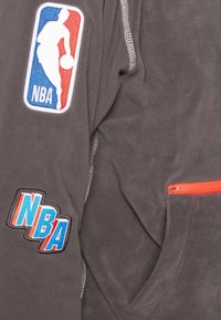 La giacca in pile grigia presenta toppe con il logo NBA in rosso, blu e bianco. Include una tasca con zip con dettagli arancioni. Tessuto morbido, design sportivo.