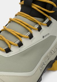 GORE-TEX μποτάκι πεζοπορίας σε ανοιχτό γκρι με μαύρες λεπτομέρειες και κίτρινες κορδόνες. Διαθέτει υφή στην επιφάνεια και έντονη λεπτομέρεια λογότυπου στο πλάι.