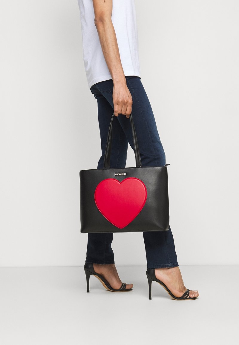 Personne portant un jean foncé et des talons noirs tenant un sac à main noir avec un grand cœur rouge dessus sur un fond blanc.
